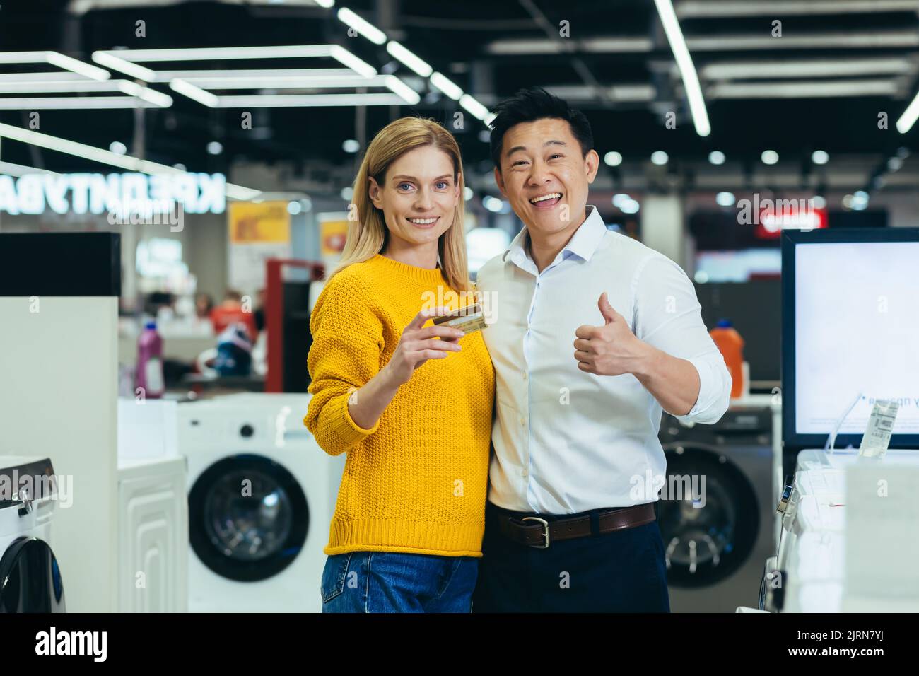 Portrait of diverse family couple Asian man and woman shopping in supermarket, electronics and household appliances, looking at camera and smiling, satisfied with bank credit card Stock Photo