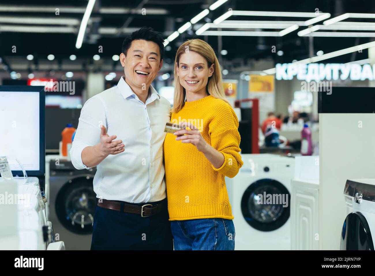 Portrait of diverse family couple Asian man and woman shopping in supermarket, electronics and household appliances, looking at camera and smiling, satisfied with bank credit card Stock Photo