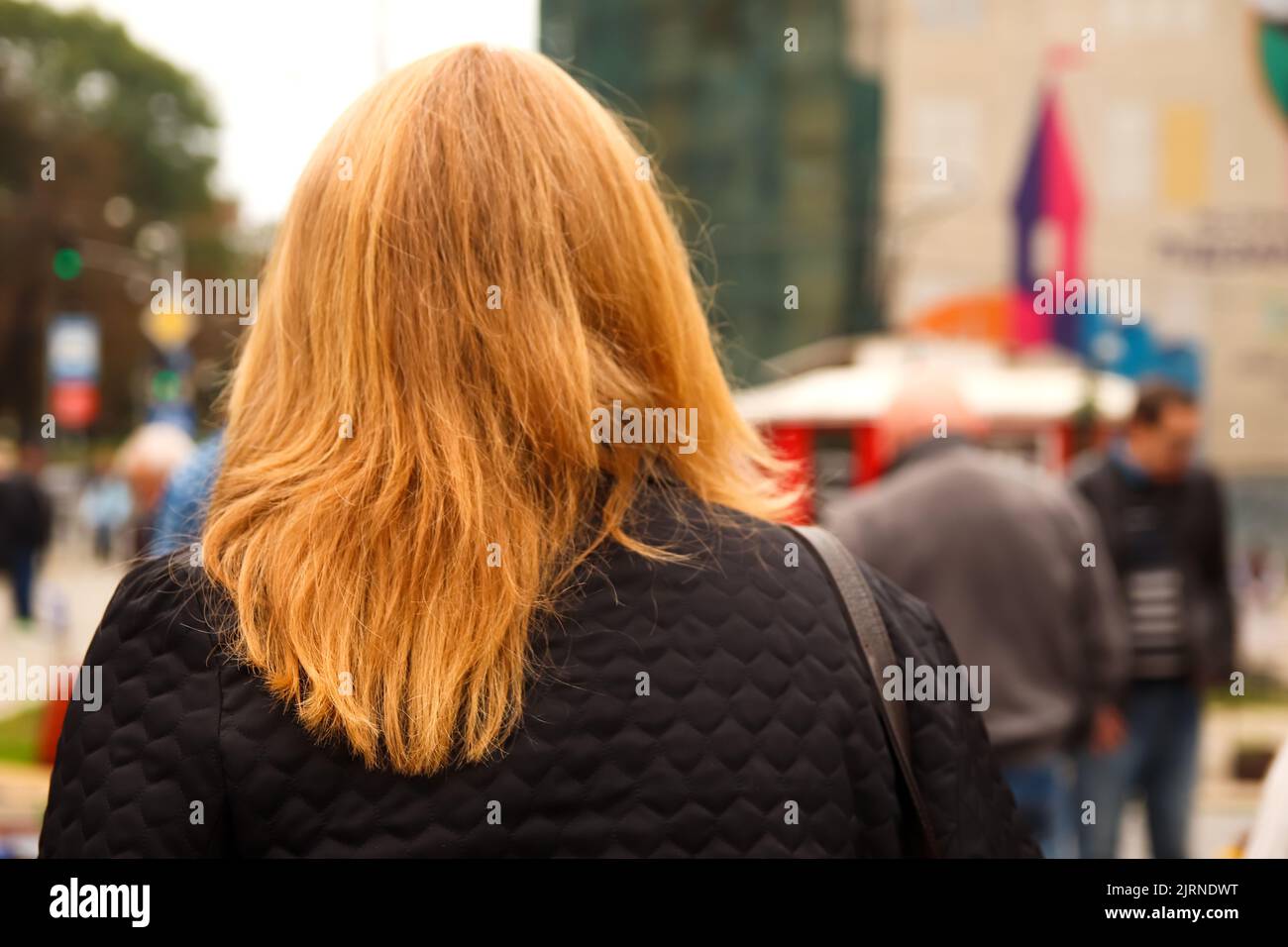 Defocus urban citizen concept. Rear view of blonde woman crossing street in city, with commuters and commercial buildings in background. Closeup one p Stock Photo