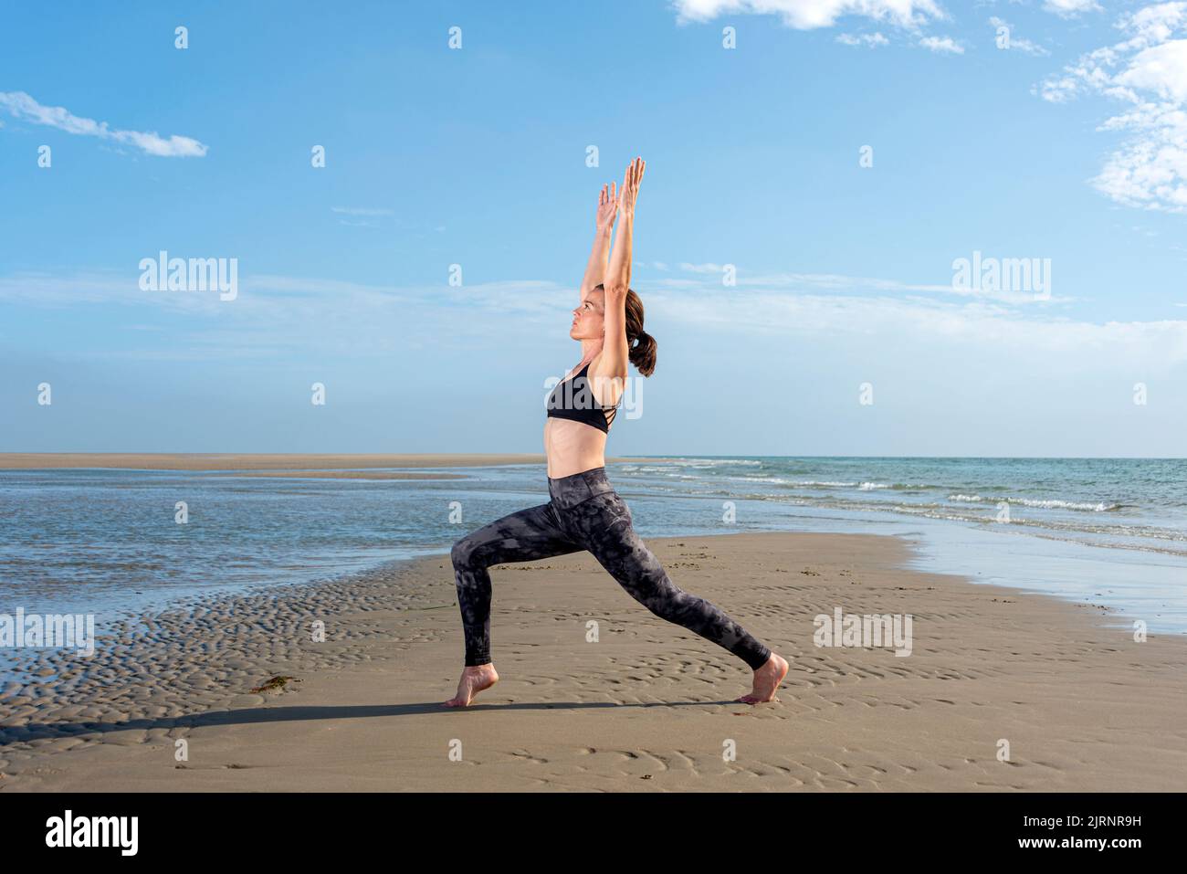 Sporty woman doing a high lunge yoga pose on a beach in the sun Stock Photo