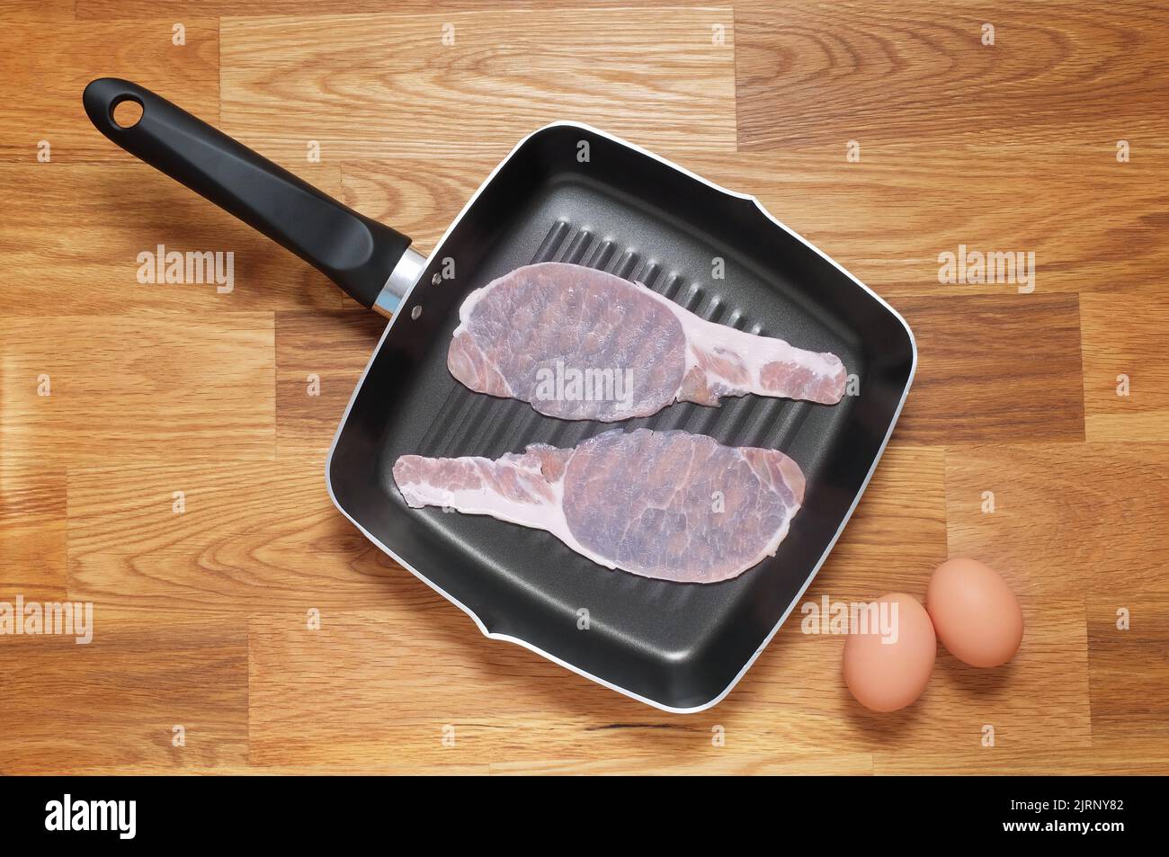 Rashers of raw bacon placed in a griddle frying pan, two brown hens eggs placed at the side on a wooden table top. Shot from above Stock Photo