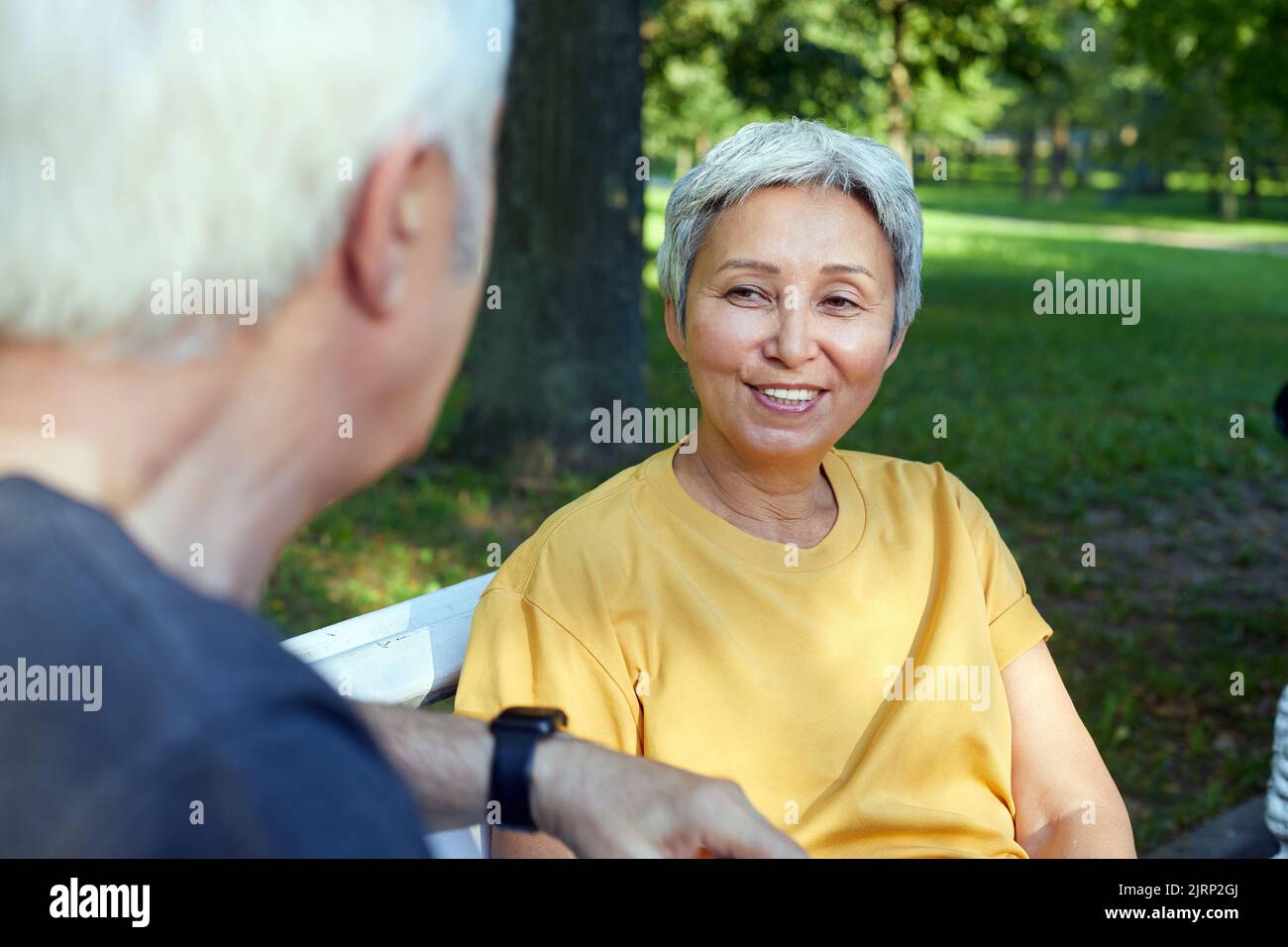Attractive older couple talking seated on bench in summer park, wear sportive clothes, enjoy conversation in the morning outdoors. Communication, harm Stock Photo