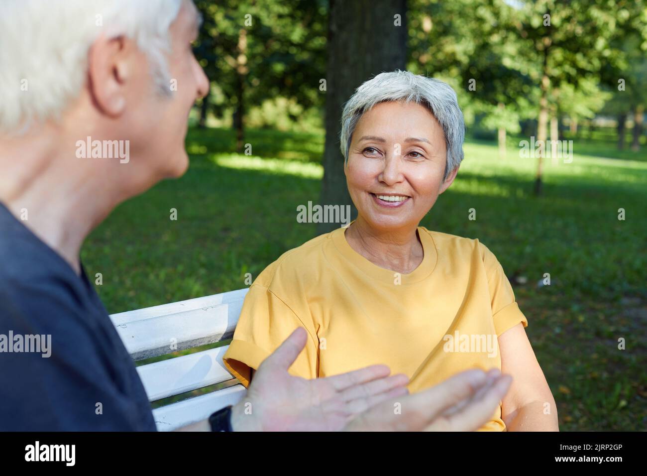 Happy older couple talk sit on bench in summer park, wear sportive clothes, enjoy conversation in the morning outdoors, focus on wife listen husband. Stock Photo