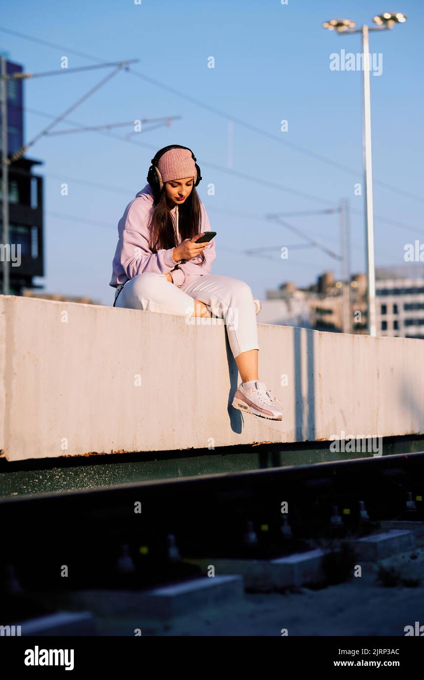 A teenage girl in urban wear sits on a railroad platform with headphones on and browses for a song on her phone. Stock Photo
