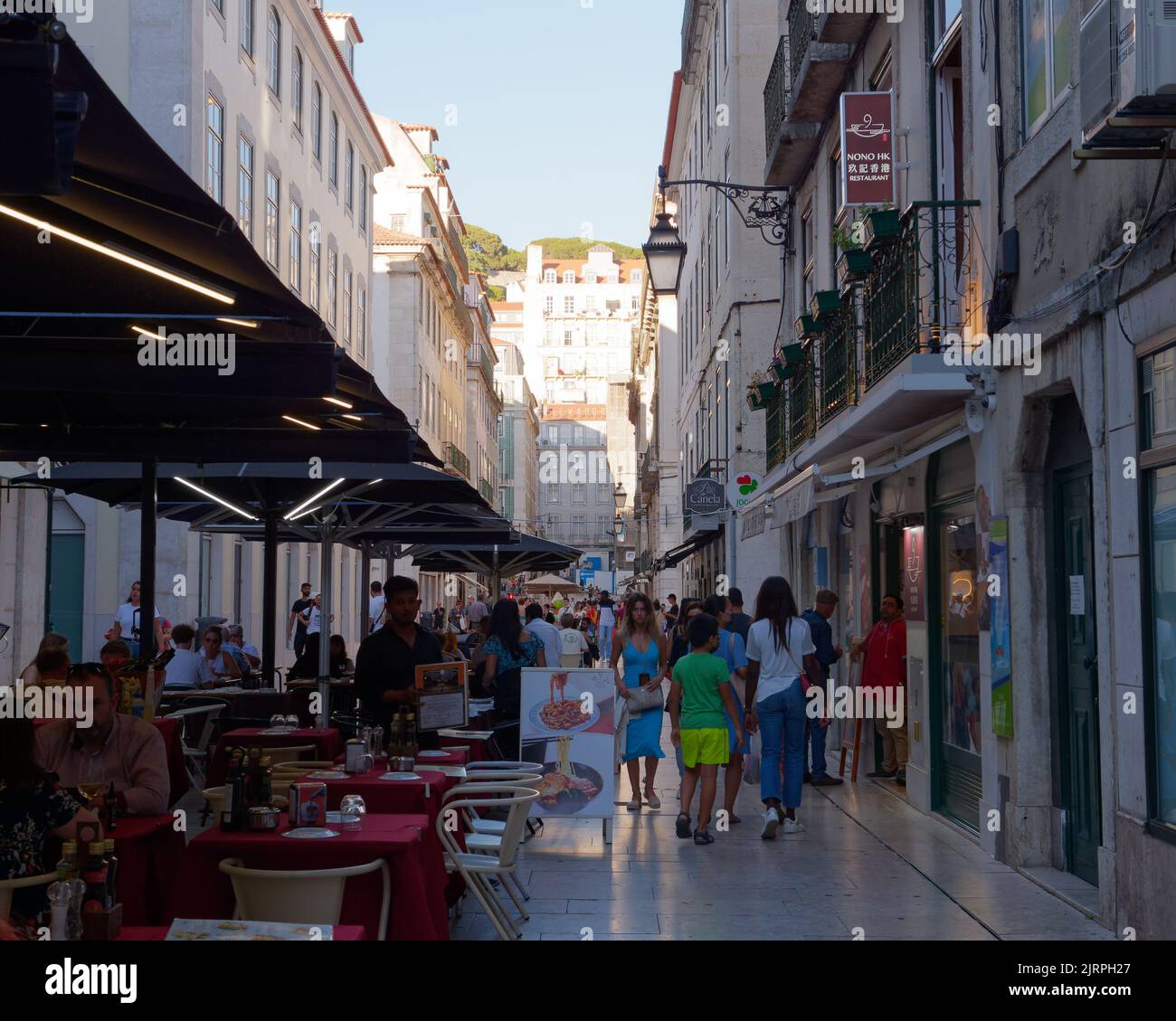 Streets of Lisbon with outside restaurants and shops with tourists browsing on a summers evening. Buildings on a hill in the background. Stock Photo