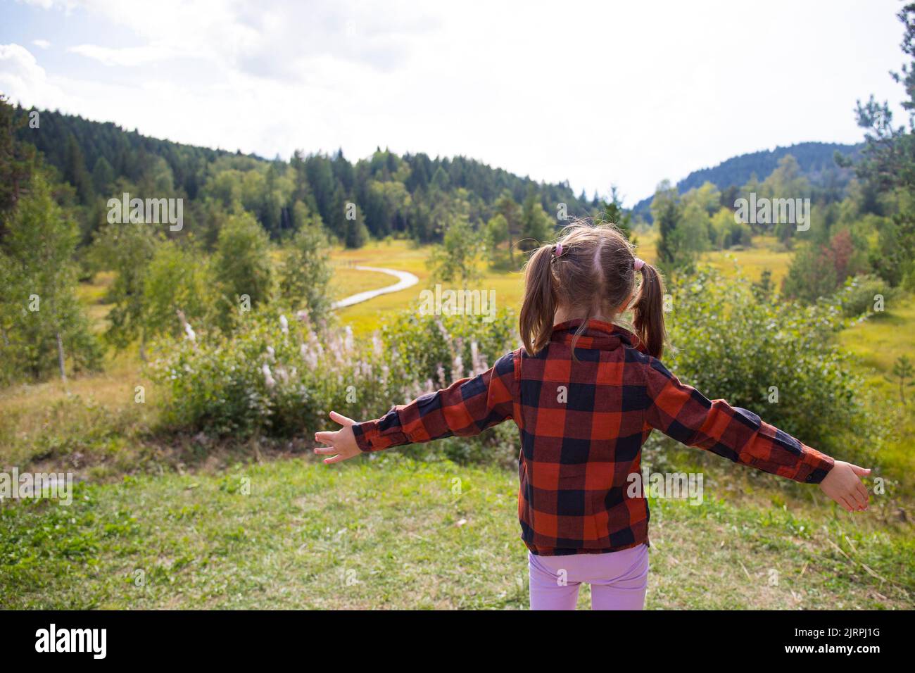 Caucasian child girl, back view, with open arms in a natural mountain landscape on a sunny day. Pian di Gembro natural park, Valtellina, Italy. Stock Photo