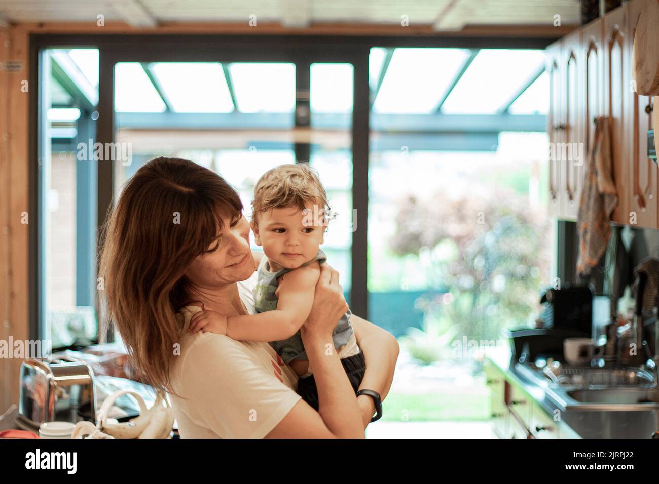 Young caucasian mother holding baby son in the kitchen. Happy simple moment, copy space. Stock Photo