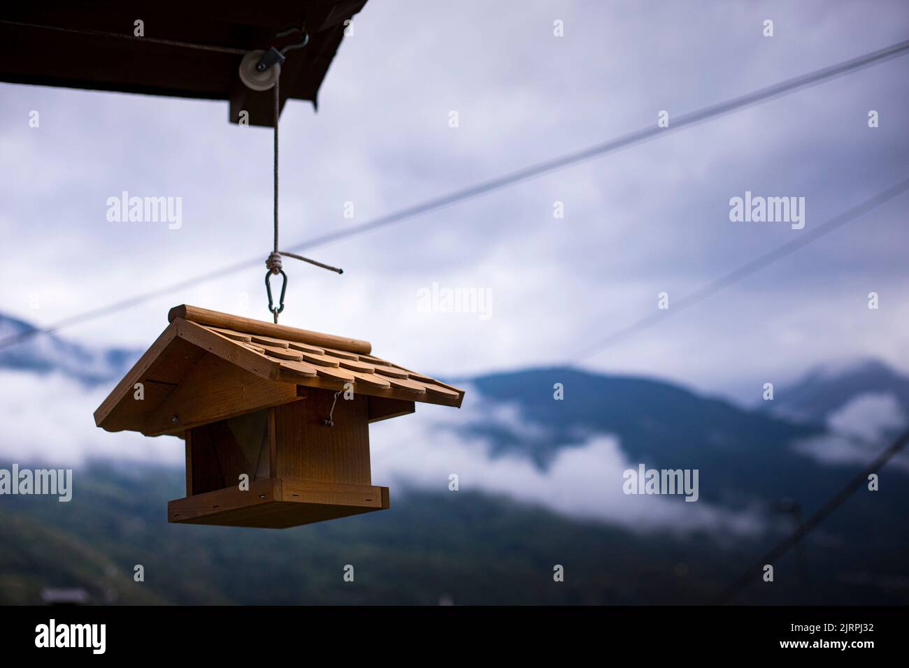 Wood brown birdhouse hanging from a roof with mountains and winter sky in the background. Wallpaper with copy space. Garden ecology idea. Stock Photo