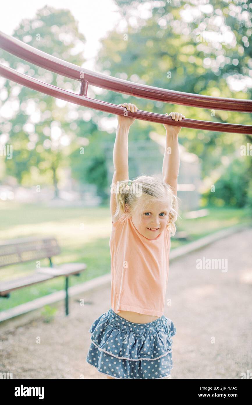 Blonde 4 year old in pigtails hangs from playground equipment. Stock Photo
