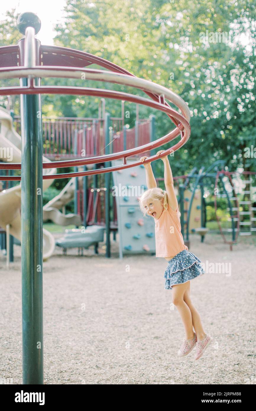 Blonde 4 year old swings from playground equipment and smiles Stock Photo