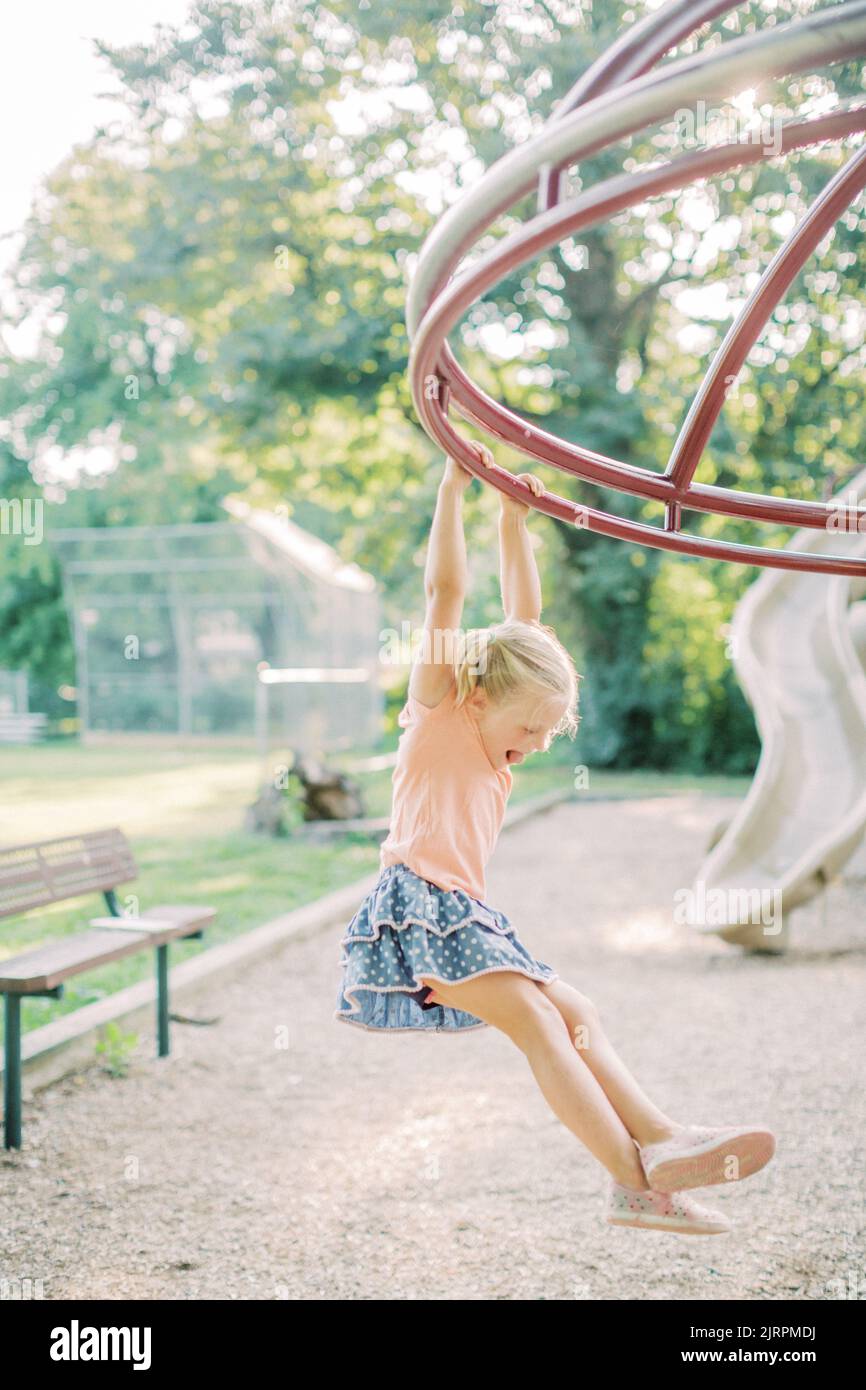 Blonde 4-5 year old girl hangs from playground equipment at sunset. Stock Photo