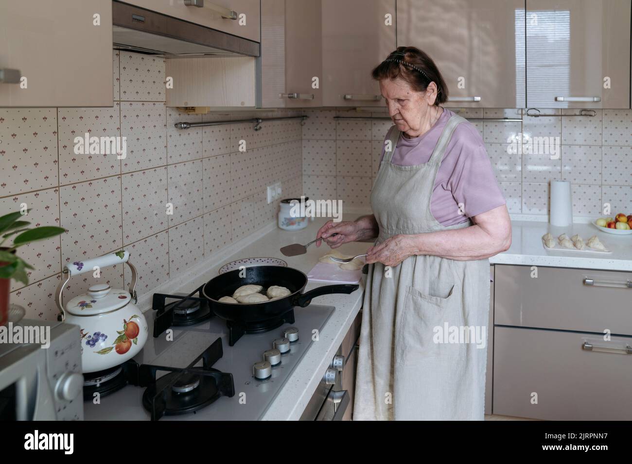 An elderly woman is frying pies in her kitchen. Stock Photo