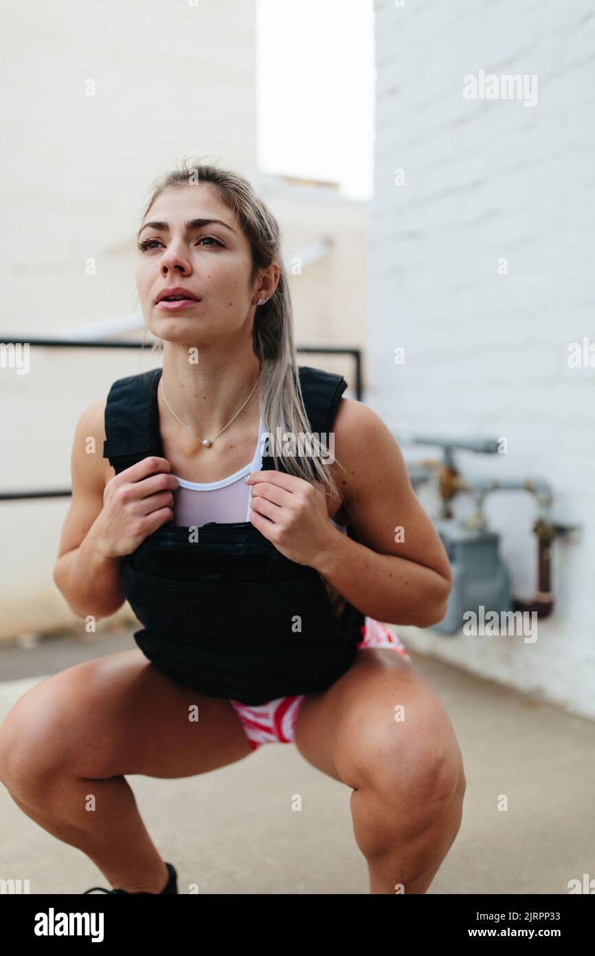 Fitness woman doing squats outside wearing weighted vest in fron Stock