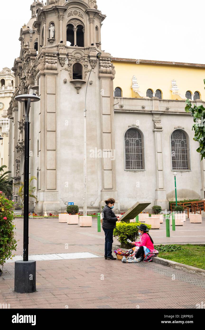 Photo of an indigenous woman selling goods on the street to another woman at Parque Kennedy, with the Catholic Cathedral Parroquiia La Virgen Milagro Stock Photo