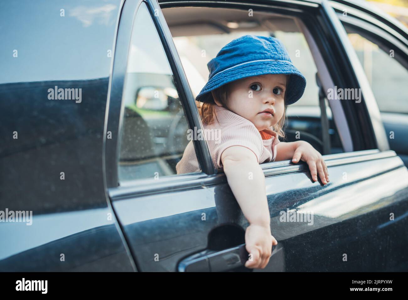 Baby girl waring hat, sticking her head out the car window and looking behind her at the camera waiting for her parents for a family trip Stock Photo