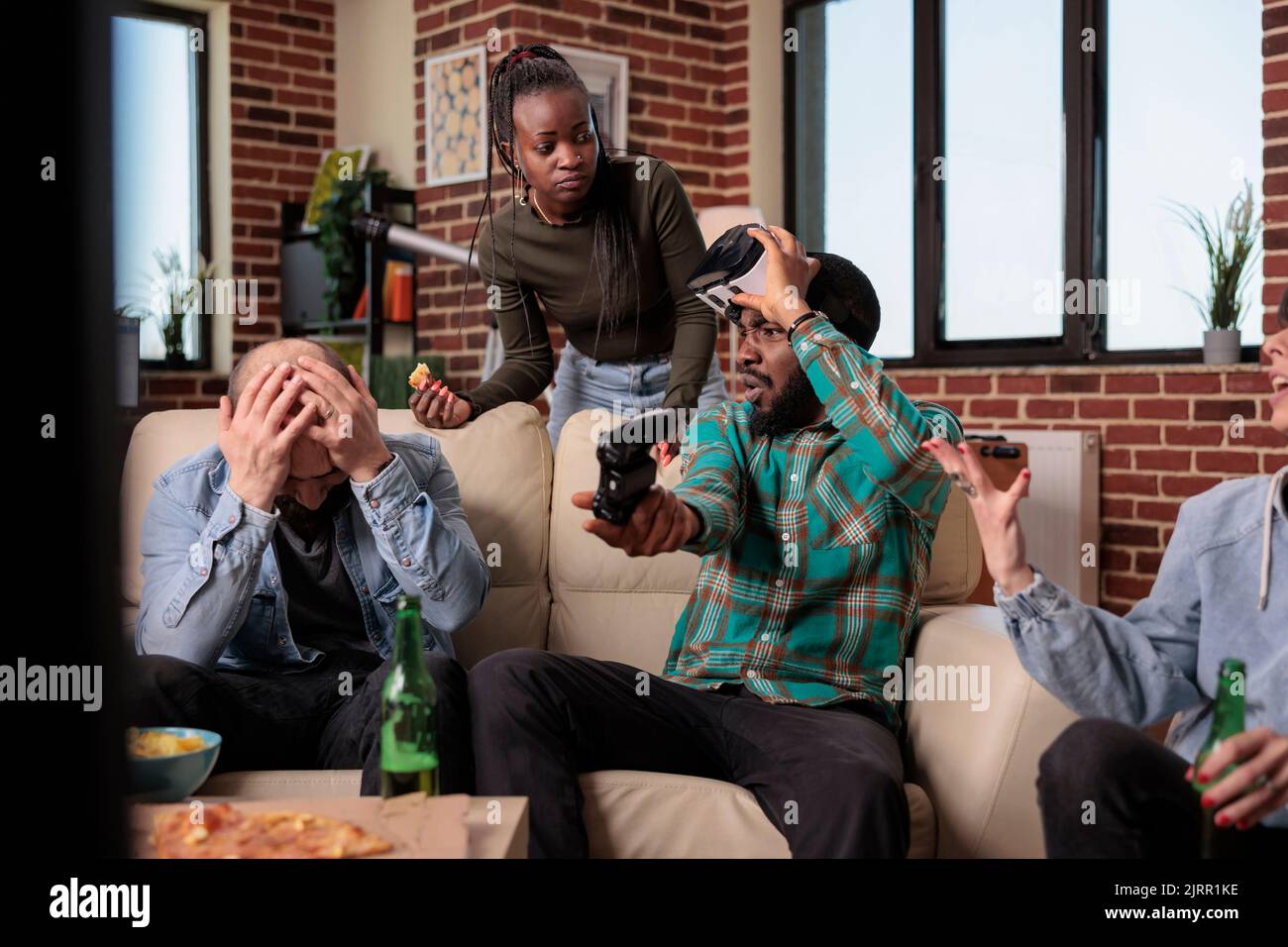Group of frustrated friends losing video games play with vr glasses and console, drinking bottles of beer. Friends feeling sad about lost gameplay competition, having fun together at gathering. Stock Photo
