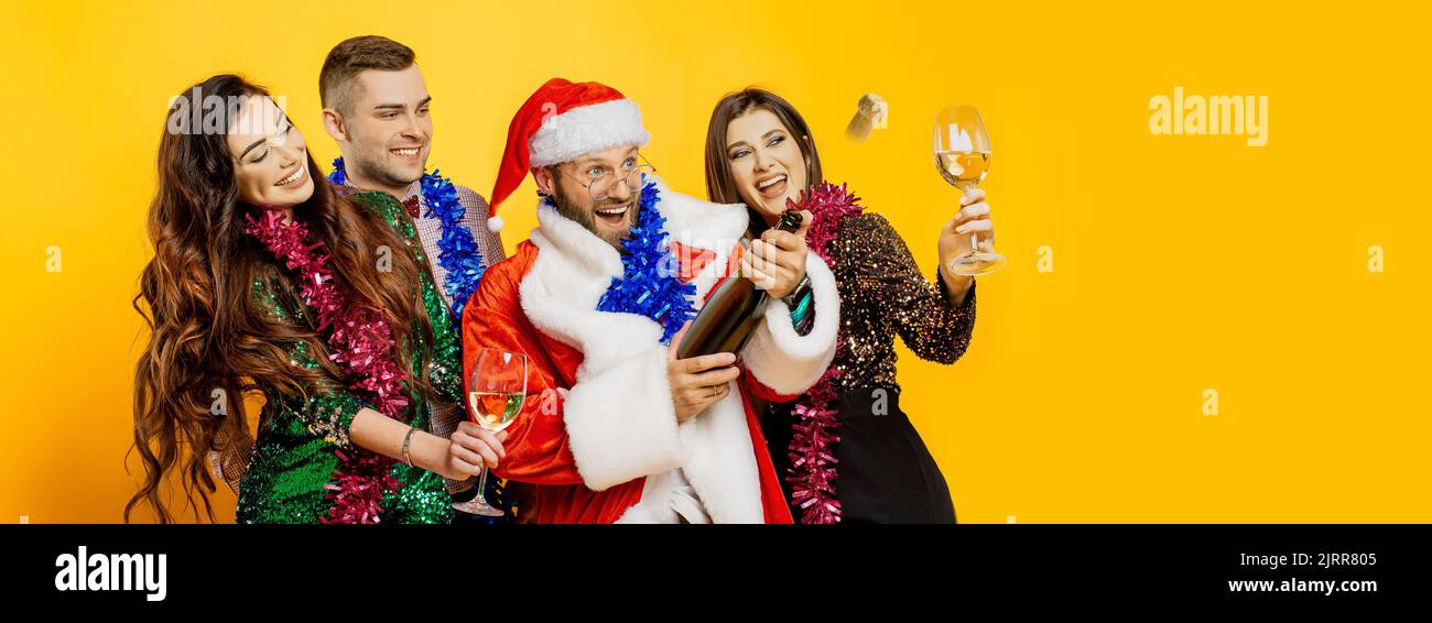 A group of young adult friends in New Year's tinsel and a santa costume shoot a champagne cork on a yellow isolated background with copy space. Banner Stock Photo