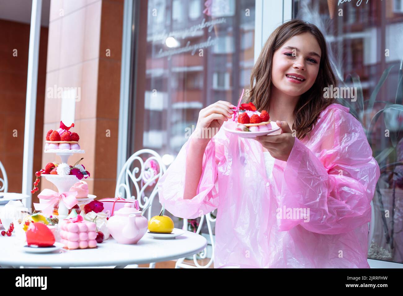 Smiling teen girl in raincoat eating sweet berry dessert cake and drink tea from table in outdoor cafe on birthday party Stock Photo