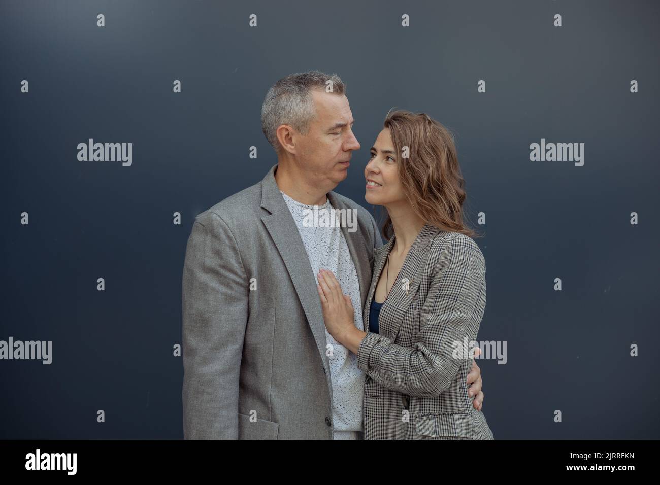 Young loving woman and handsome aged gray haired partner embracing and looking to each other, gray background. Romantic relations of beautiful couple Stock Photo
