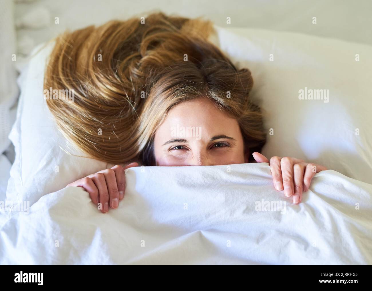 Sunday is duvet day. Portrait of a playful young woman hiding under the covers in her bed. Stock Photo