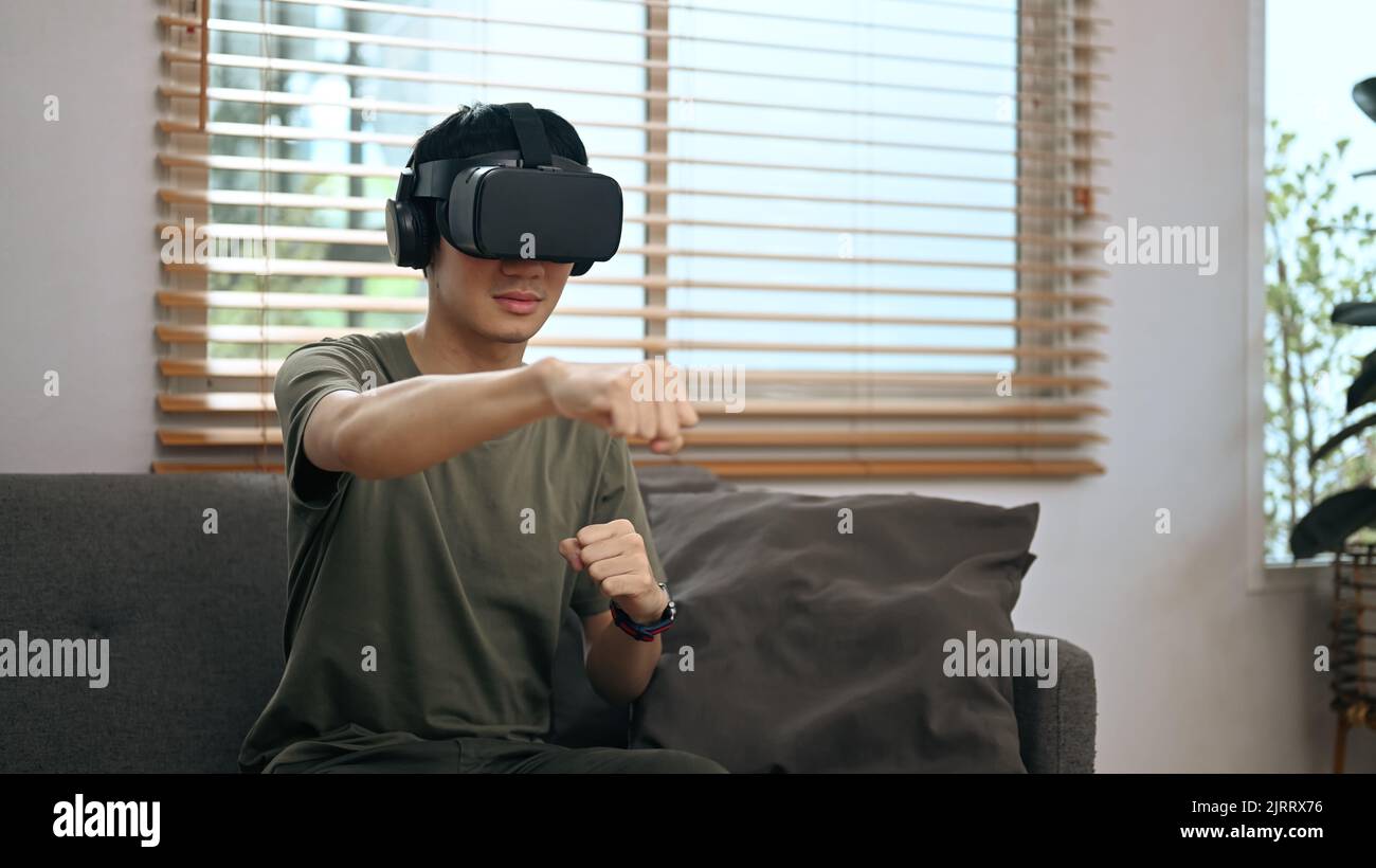 Happy man wearing virtual reality headset playing simulation boxing game at home. Future