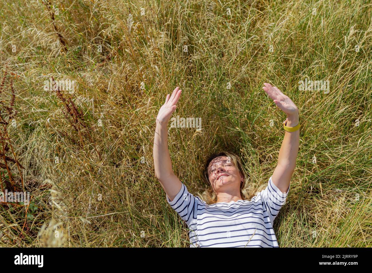 A woman lies on the grass and happily reaches out with her hands to the sun. Stock Photo