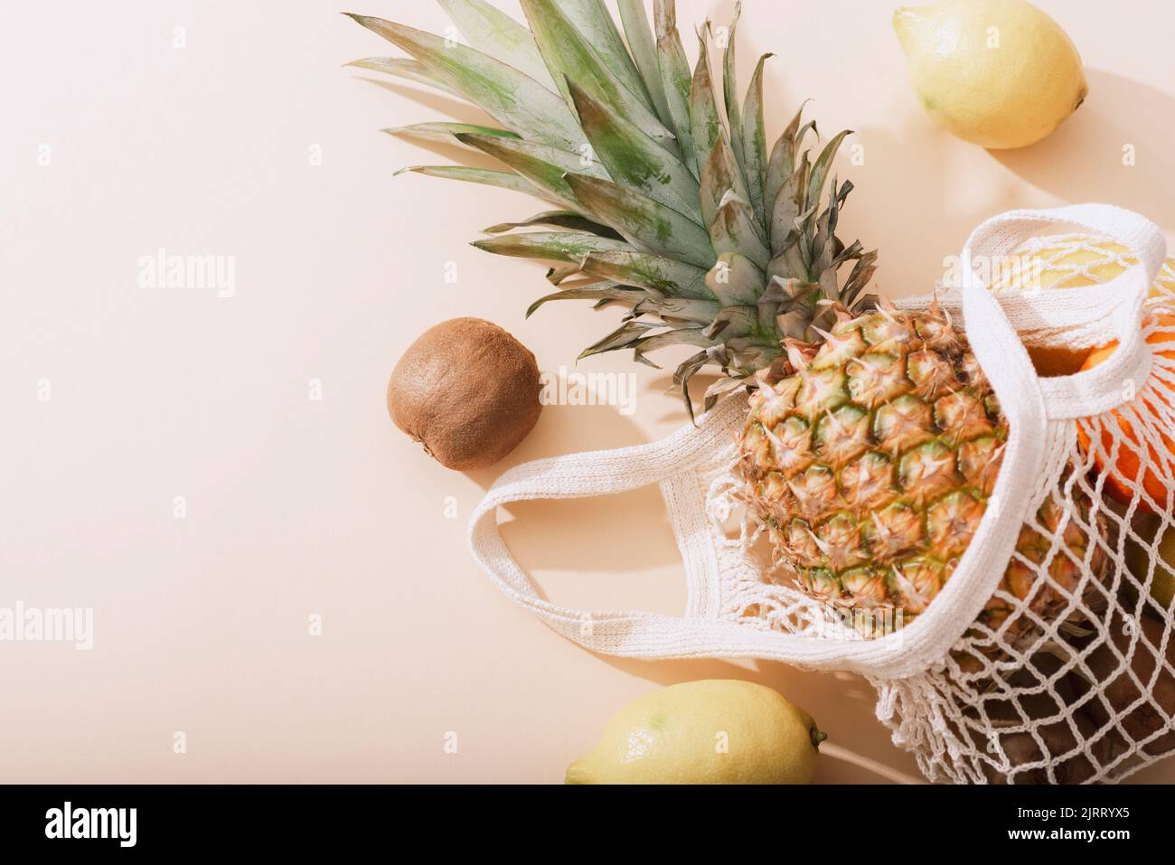 Different tropical fruits in mesh bag on beige background. Pineapple, kiwi, lemons and oranges. Top view, flat lay, copy space. Stock Photo