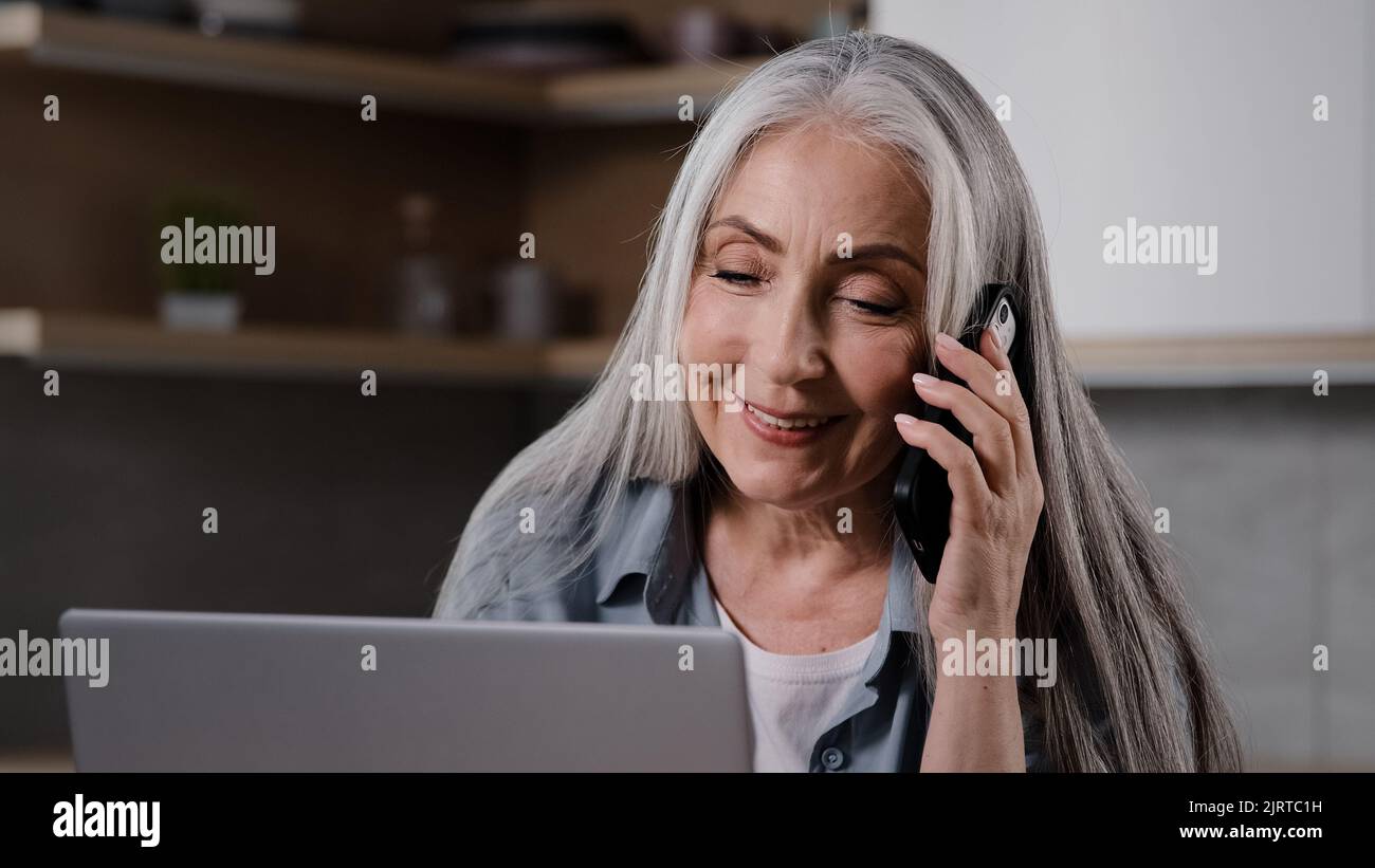 Aged business lady experienced female specialist talk mobile phone look at laptop screen negotiate with client in home kitchen. Grey-haired housewife Stock Photo