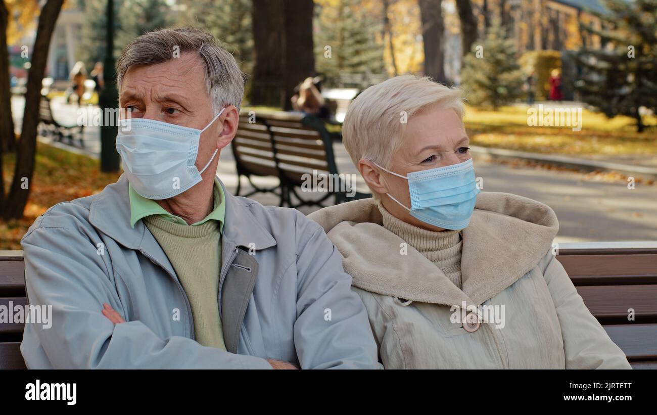 Elderly family couple in medical masks sit in autumn park mature man turns away resentfully crossing arms unpleasant conversation outdoors Stock Photo