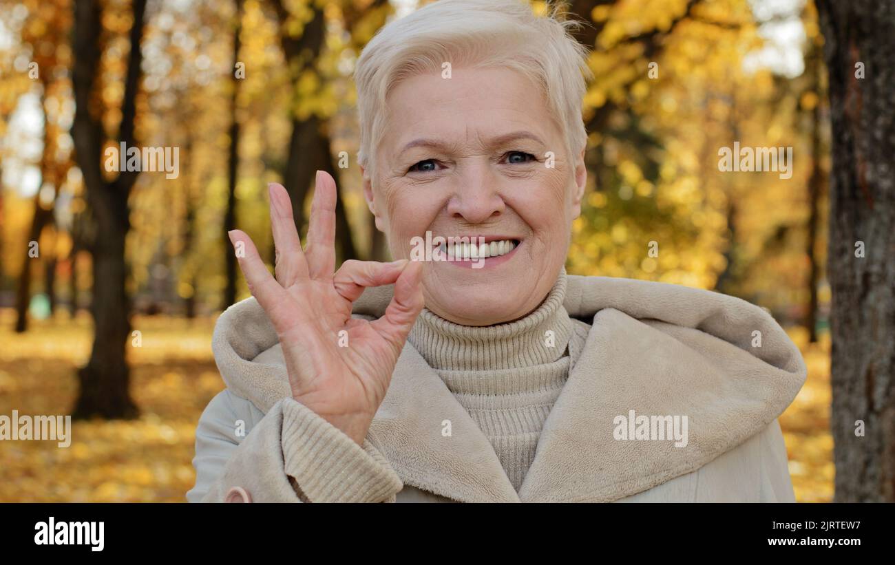 Happy elderly woman joyfully smiling standing in autumn park mature grandmother looking at camera showing gesture okay symbol of satisfaction Stock Photo