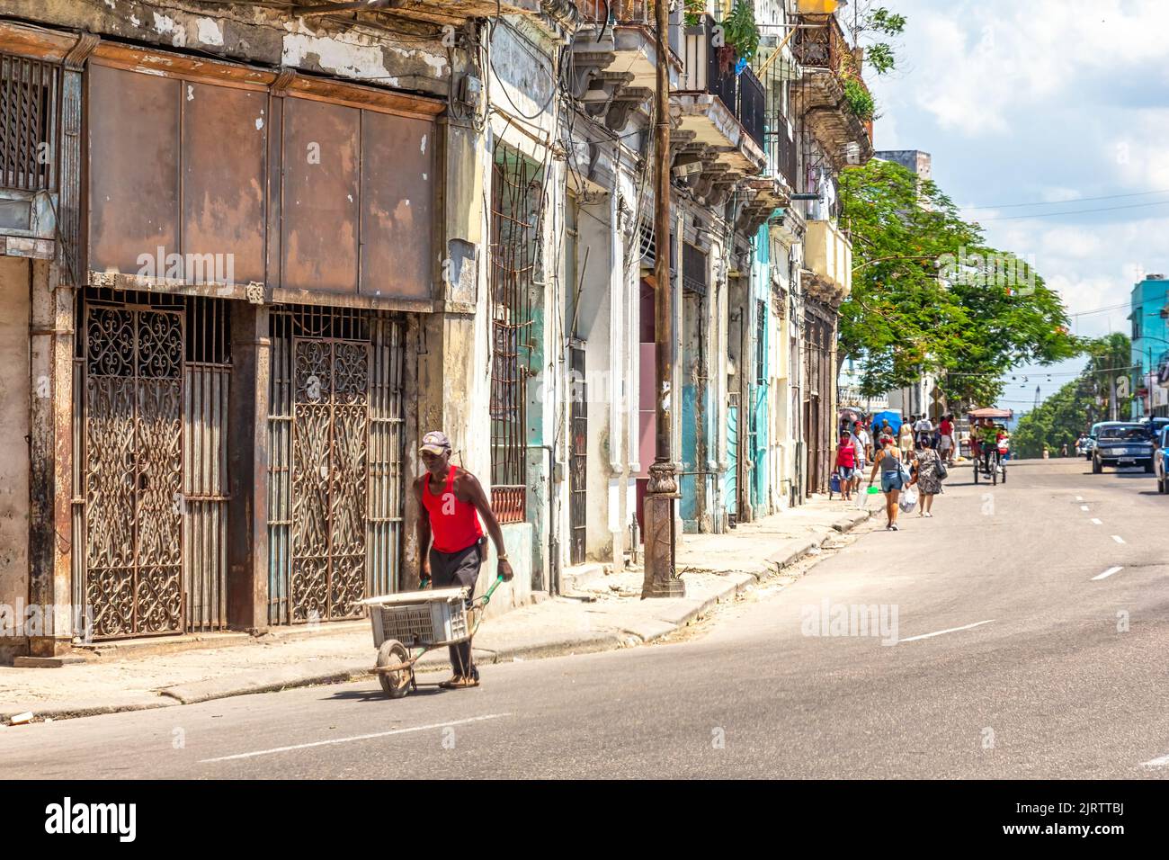 A Cuban afro-caribbean man pushes a wheelbarrow in a city street. He is passing by old, run-down, and weathered buildings Stock Photo