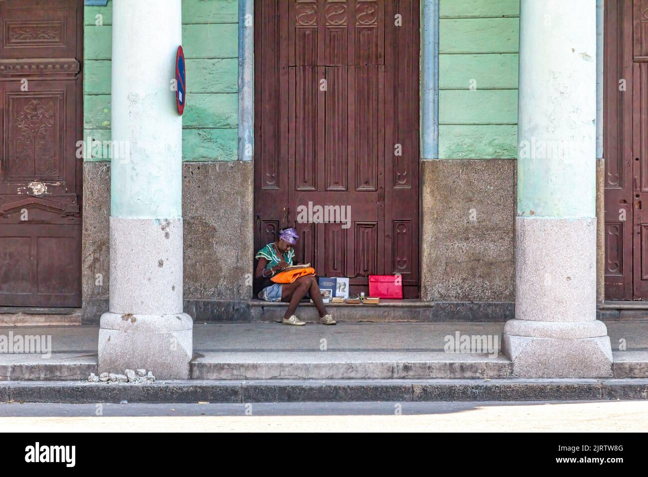 A Cuban afro-caribbean woman sits in a doorstep below a porch. She has a few books with her. Stock Photo
