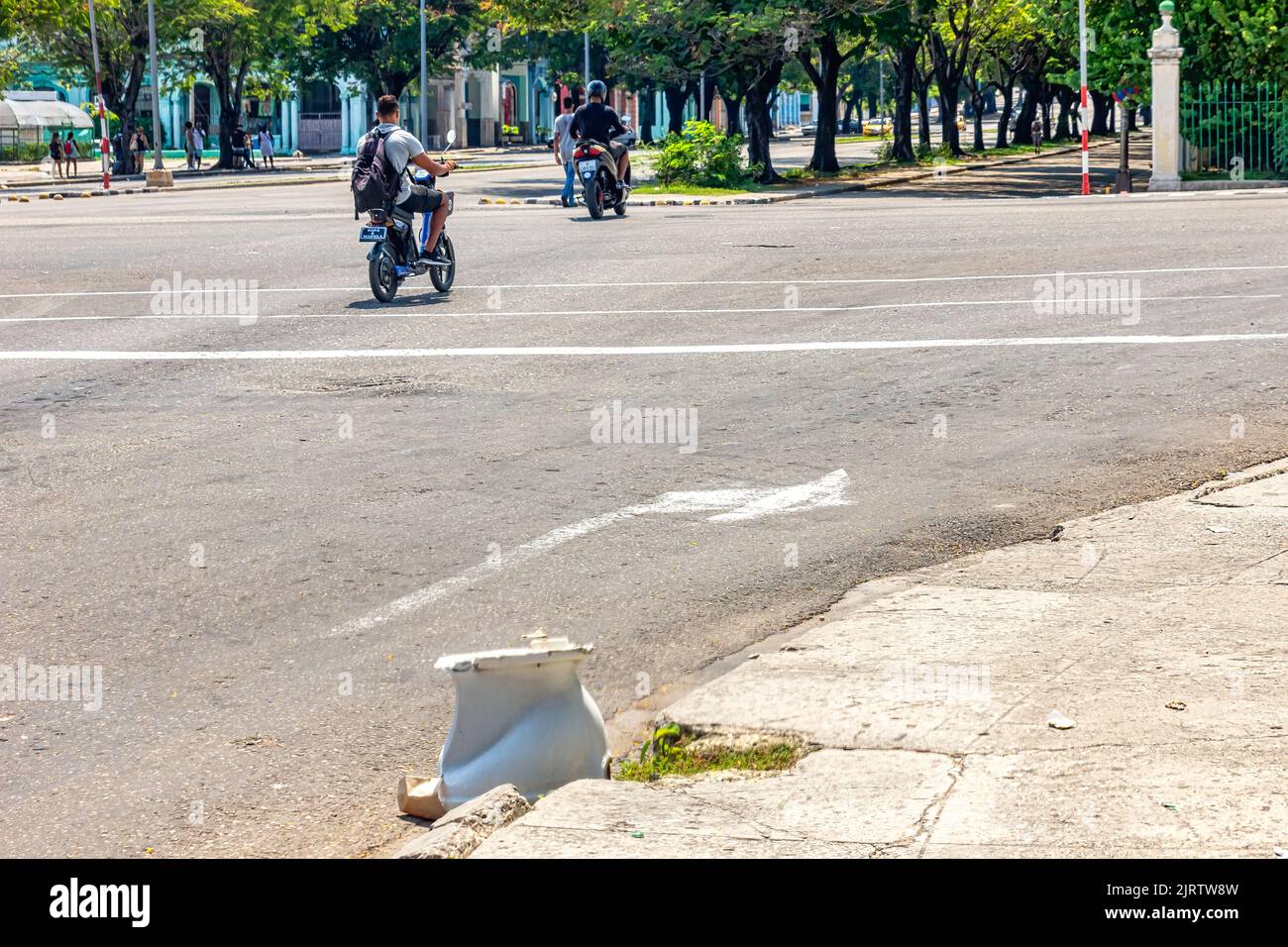 Cuban people driving motorcycles in a city street where a broken toilet   is discarded Stock Photo