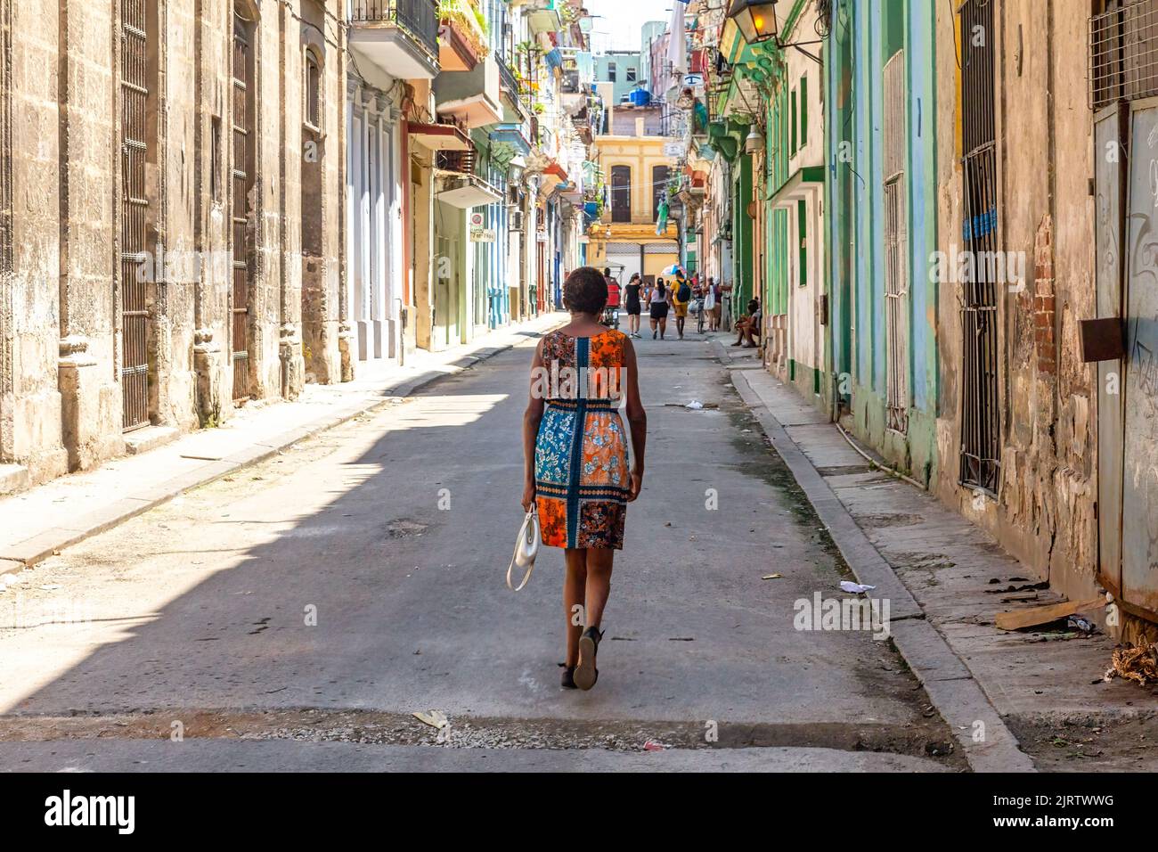 A Cuban Afro-Caribbean woman walks in a city street where a weathered facade of buildings is lined up. The street is with holes and the sidewalk is dir Stock Photo