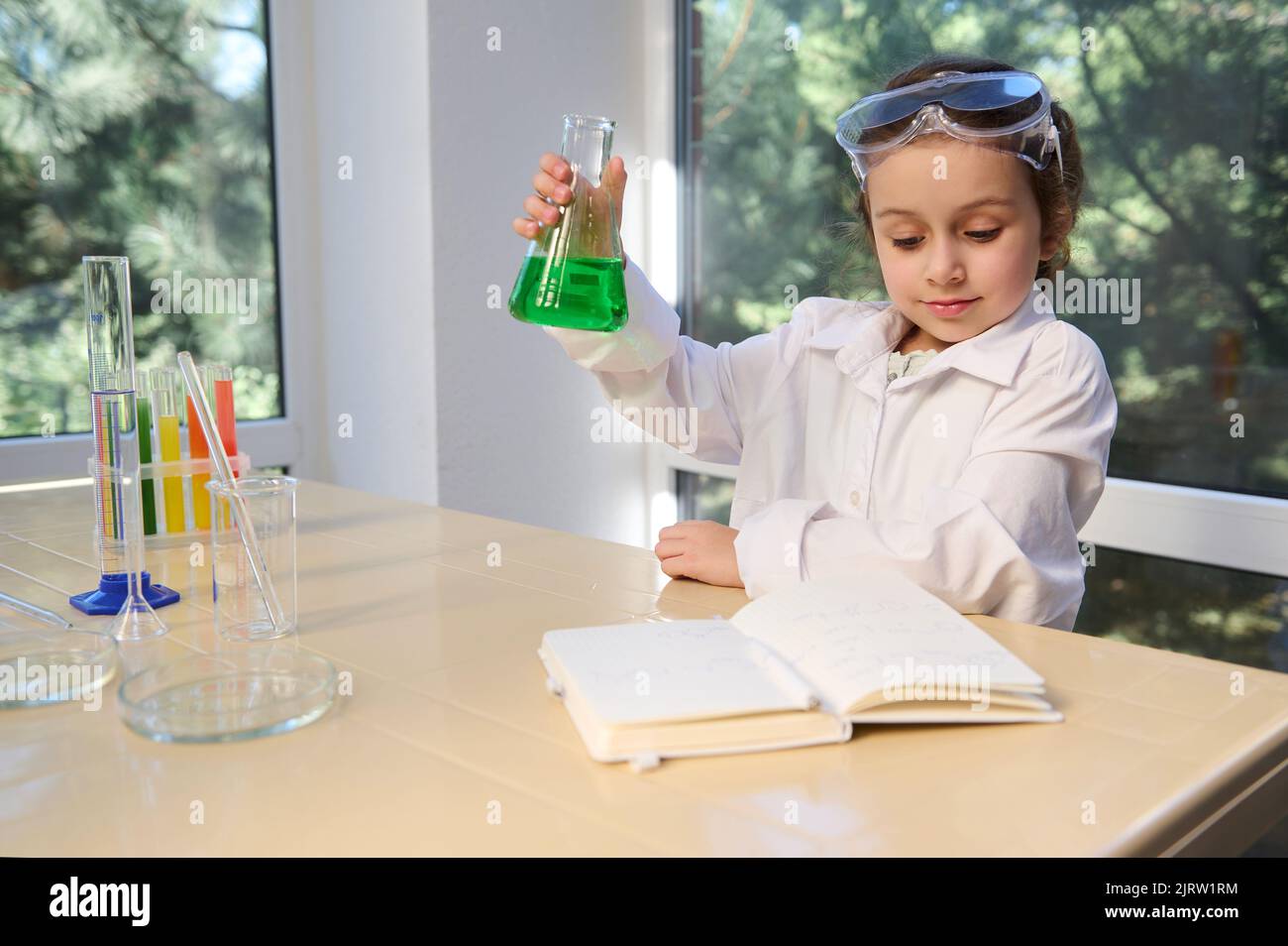 Delighted schoolgirl in white lab coat and safety goggles, testing chemistry lab experiment and