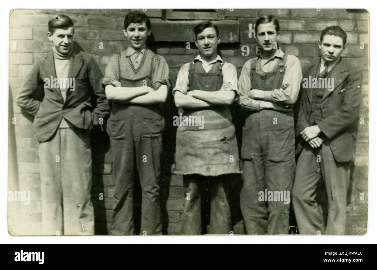 Original 1930's era postcard of five cheerful looking working class lads, possibly on apprenticeships, in industry or construction. One young man is wearing an apron, 2 are wearing overalls, 2 are wearing suit jackets and trousers, unknown location in the U.K. Stock Photo
