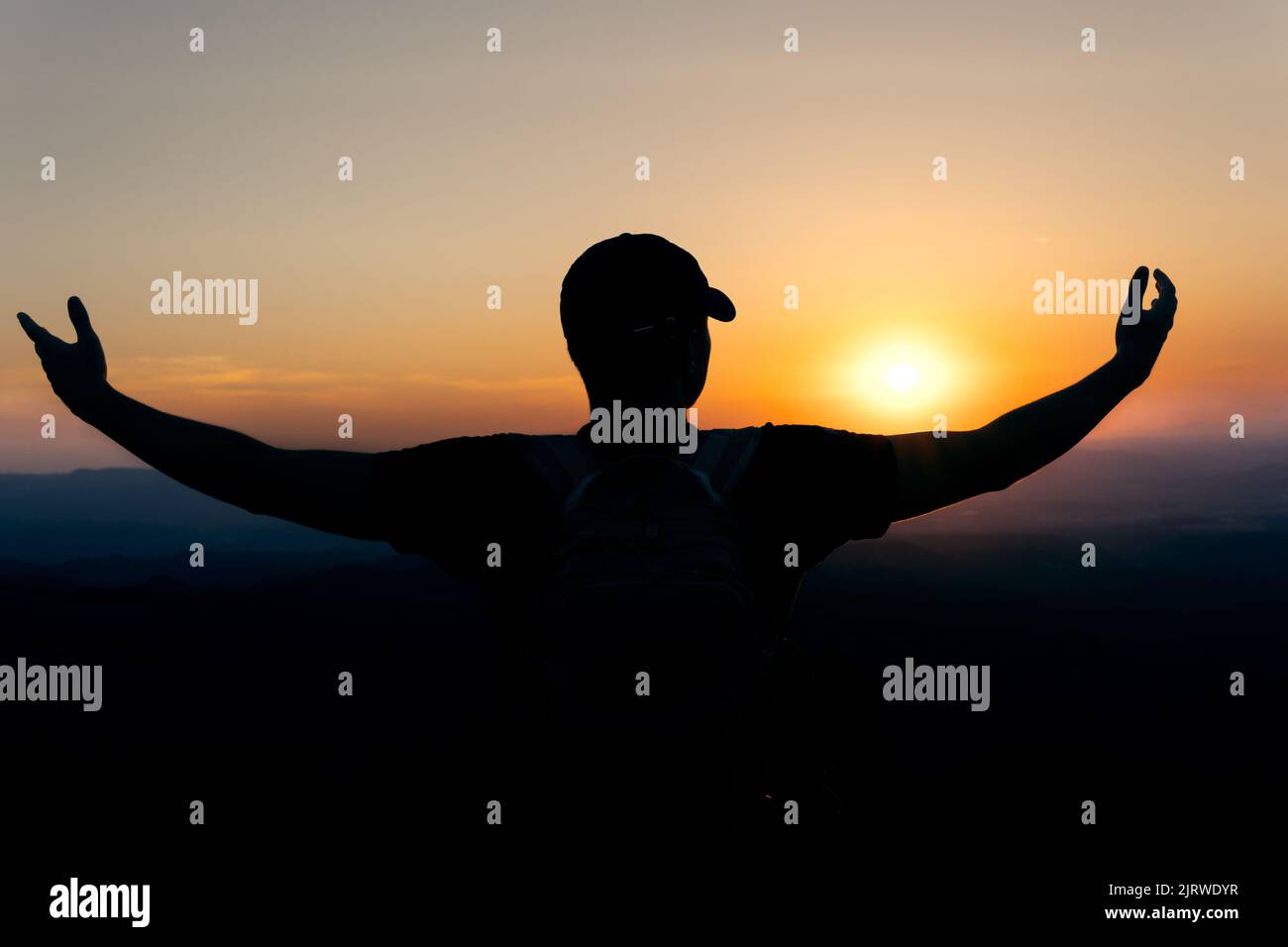 Sunset with a silhouette of a young man with a cap in an attitude of gratitude to the sun, with a background of mountains. Stock Photo