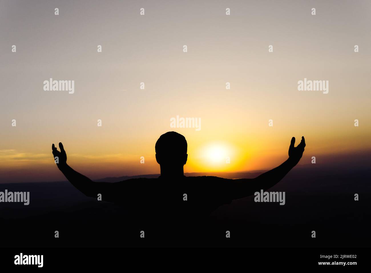 Sunset with a silhouette of a young man with a cap in an attitude of gratitude to the sun, with a background of mountains. Stock Photo