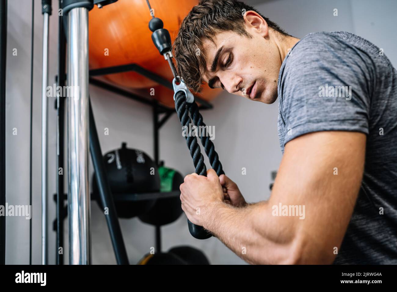 Low angle of strong male athlete with strong arm muscles looking at bicep and pulling rope of cable machine during fitness training in contemporary gy Stock Photo