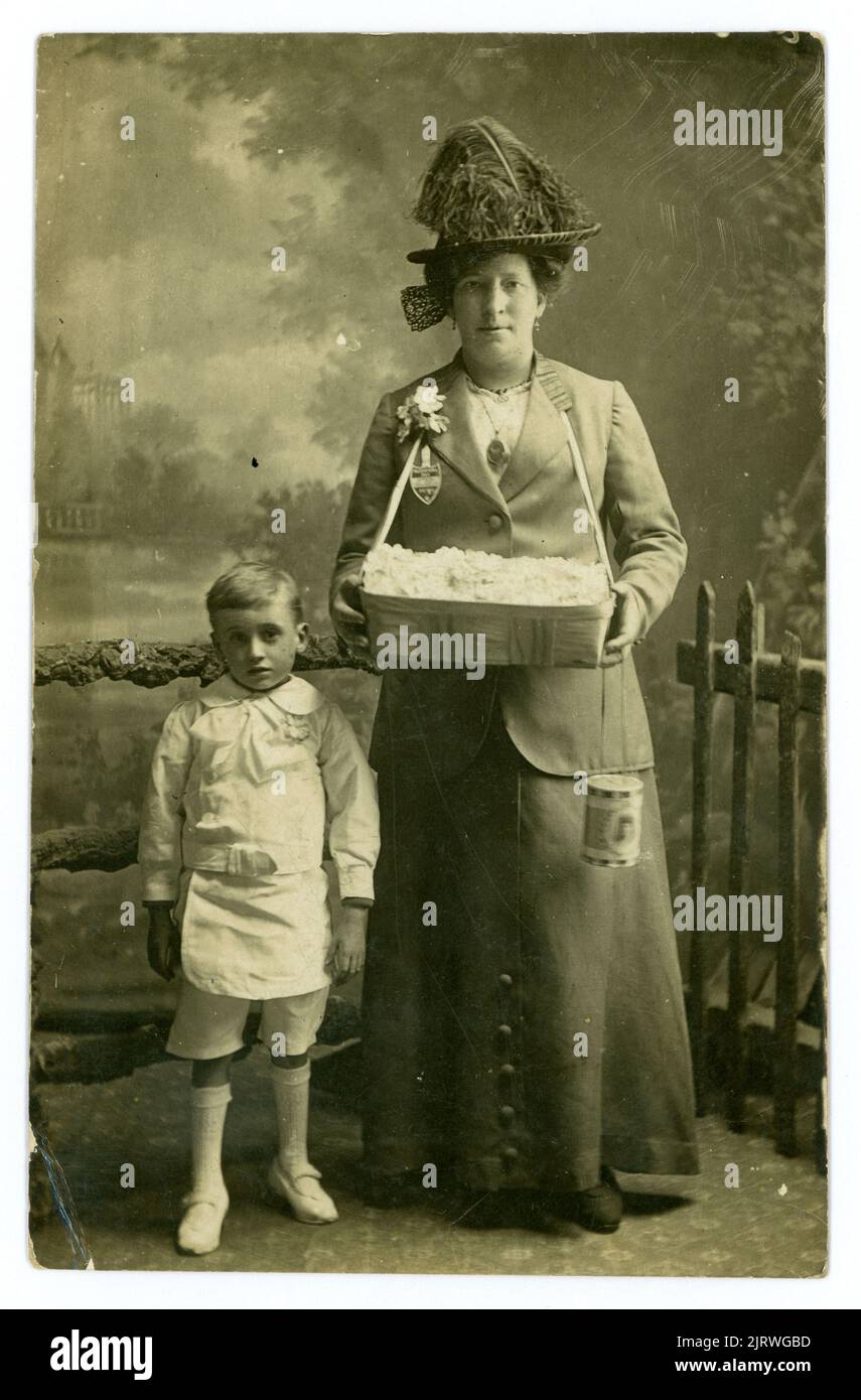 Original clear, studio portrait postcard of older, mature woman. She is an Alexandra Day charity rose seller and the child standing next to her is possibly a poor child or orphan of Bethnal Green. The child is dressed smartly but in an ill-fitting white suit and  shoes which are too small. The woman holds a collecting tin which has the patron, Queen Alexandra's portrait on it.  The woman is wearing a badge that denotes she is an official collector. From studio of  A. Lewis 228 Green St. Bethnal Green, London, U.K  circa 1913. Stock Photo