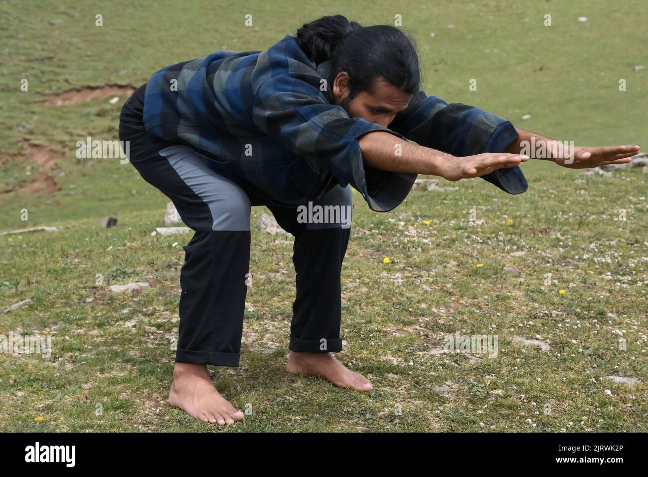 A long haired Indian young man practicing Bear Pose (Chair Pose Variation Forward Stretch) in outdoor. A young guy doing Utkatasana yoga pose (Variation Forward Stretch). Stock Photo