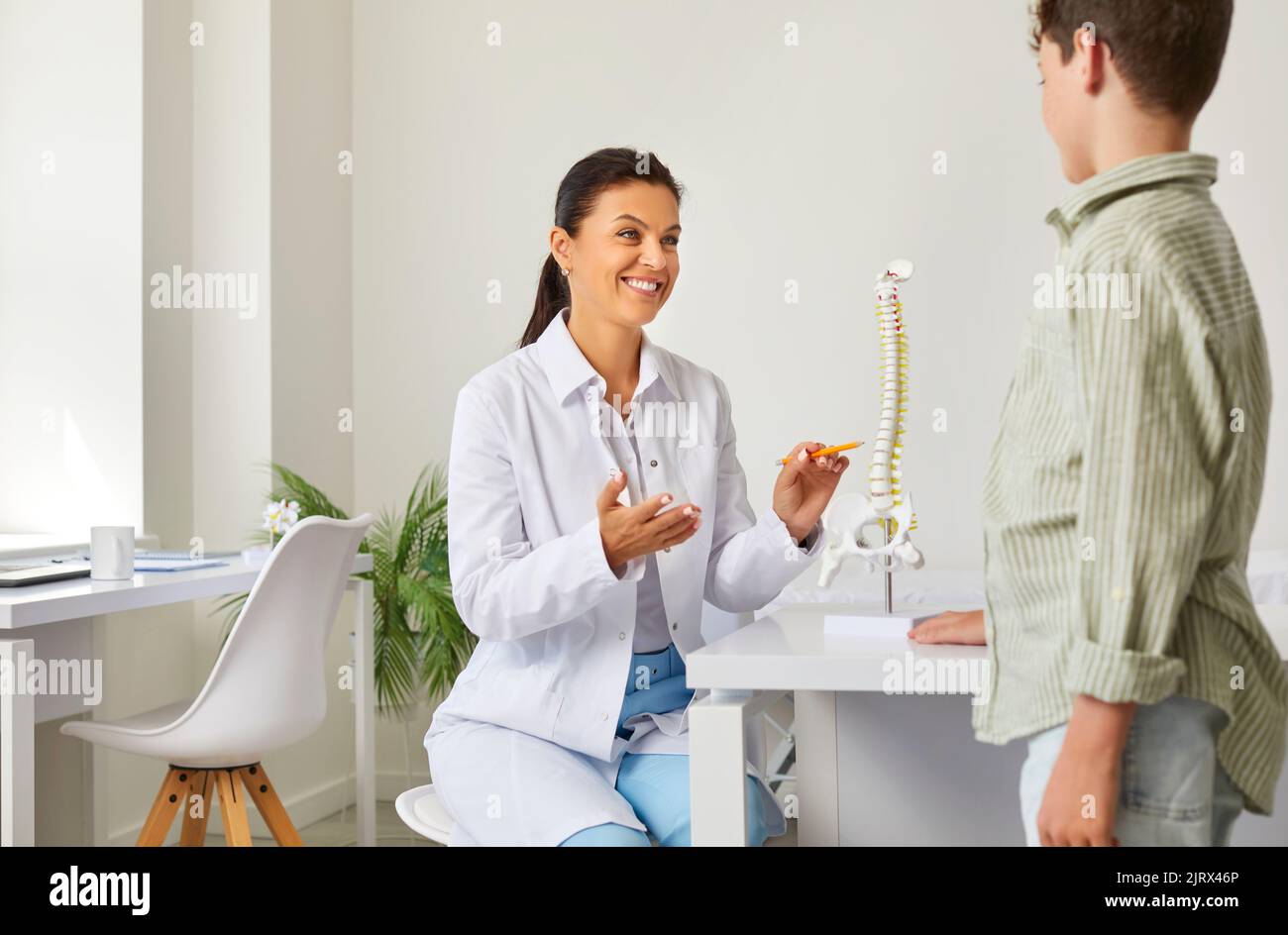 Physiotherapist tells preteen boy about structure of spine, model of which is standing on table. Stock Photo