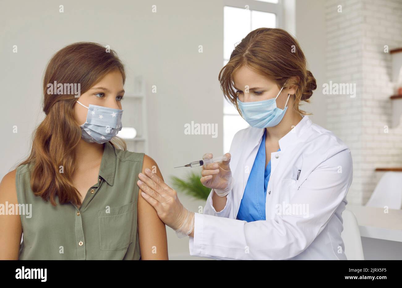 Doctor at vaccination center gives Covid 19 vaccine to teenage girl wearing face mask. Stock Photo