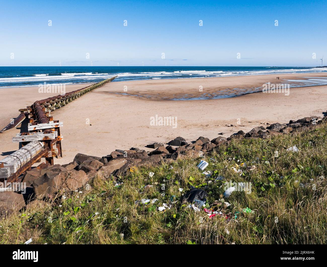 Outfall pipe at Cambois, Northumberland, UK into blue sea with wind turbines in the distance with litter in foreground. Stock Photo