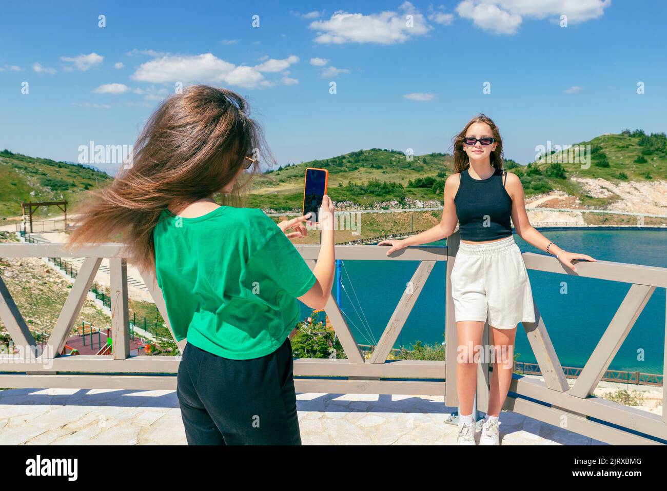 A young girl takes a photo of her friend in front of the lake with a mobile phone. The concept of travel influencers. Mountain in the background Stock Photo