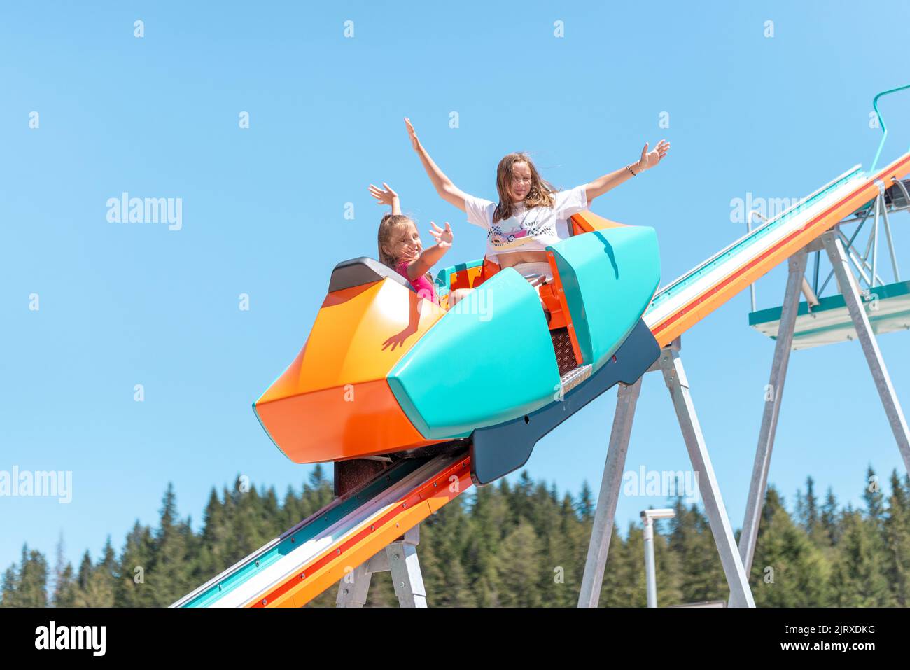 Girls are going down the hill in a mountain coaster with outstretched arms. Mountain trees in background Stock Photo