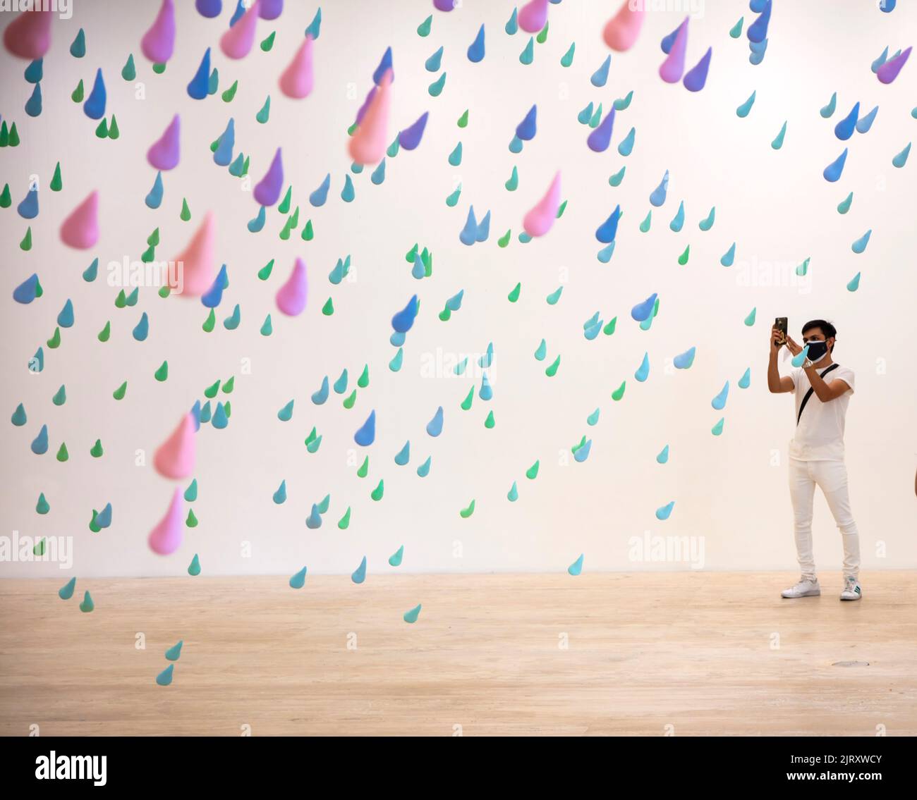Man wearing face mask takes smartphone photo of colourfull rainrops art installation by Swiss artist Urs Fischer in the Jumex Museum, Mexico City, Mex Stock Photo