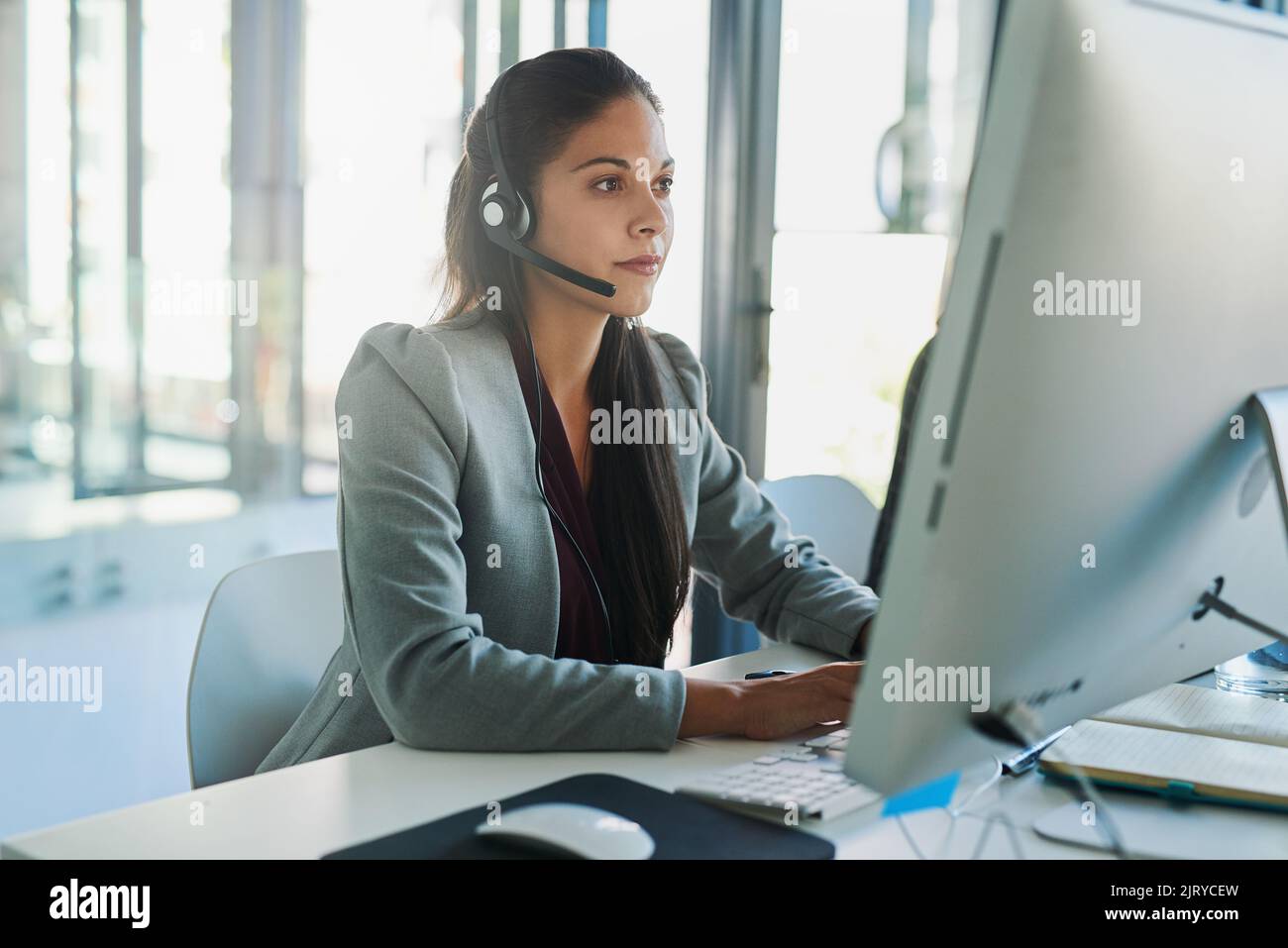 She has a strong client focus. a young woman wearing a headset and using a computer at her work desk. Stock Photo