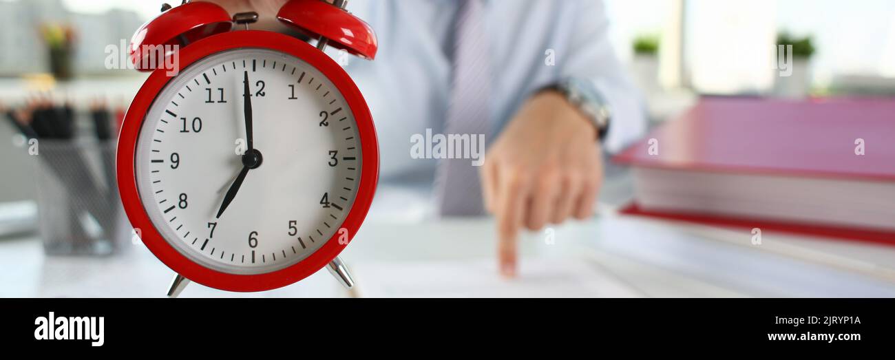 Man hand on red alarm clock stands at desk in office showing seven o