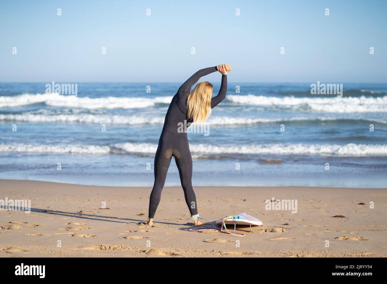 Surfer warming up at the beach with her surfboard on her side in the morning before surf. Stock Photo